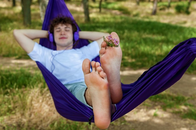 A young guy lying in a purple hammock listens to music on headphones while enjoying nature