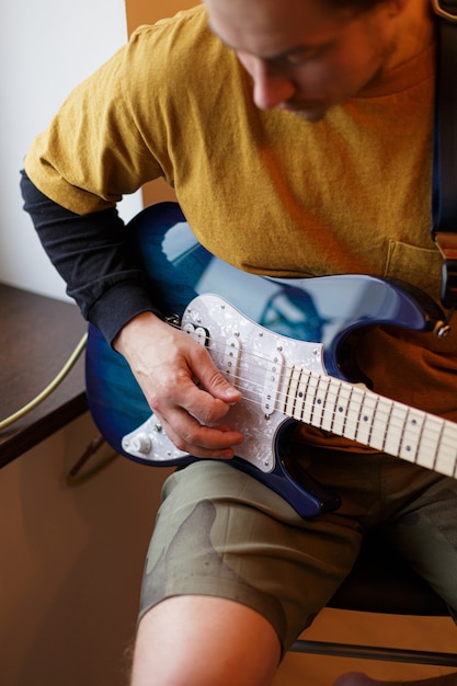 A young guy plays an electric guitar sitting by the window