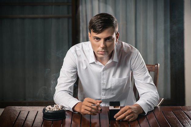 Young guy posing sitting at a table on which stands an ashtray full of cigarettes