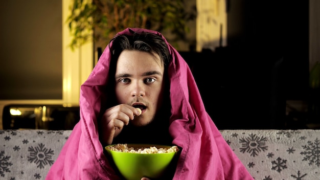 A young guy in a red T-shirt is sitting on the couch under a pink blanket, eating popcorn and watching TV intently.