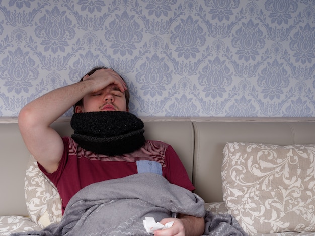 Young guy in a red t-shirt sits on the bed and takes cough pills, pain relievers and cold medications, the concept of illness at home