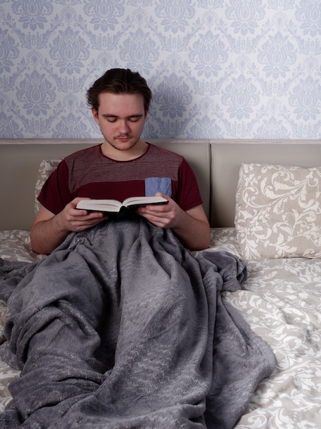 A young guy in a red T-shirt sits on a light-colored bed and reads a thick book, the concept of rest after a working day