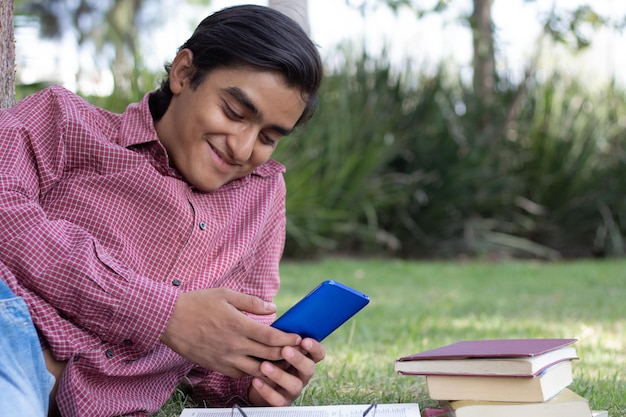 Young guy relaxing in the park reading books and looking at his cell phone screen laughing