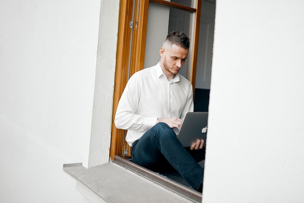 A young guy sits on the windowsill at home and works with a laptop