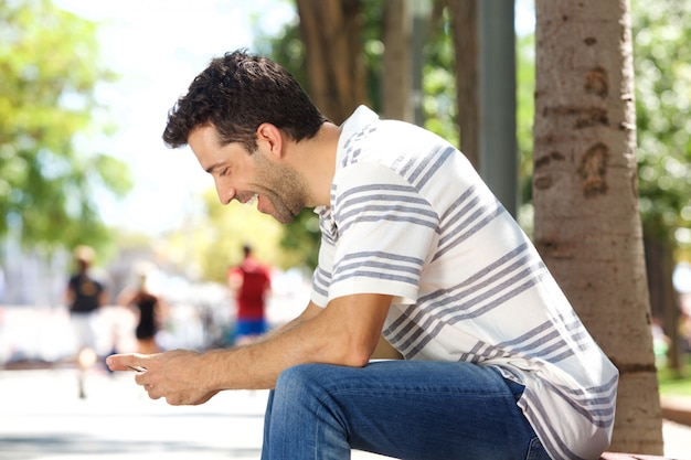 Young guy sitting outdoors smiling and sending text message on mobile phone