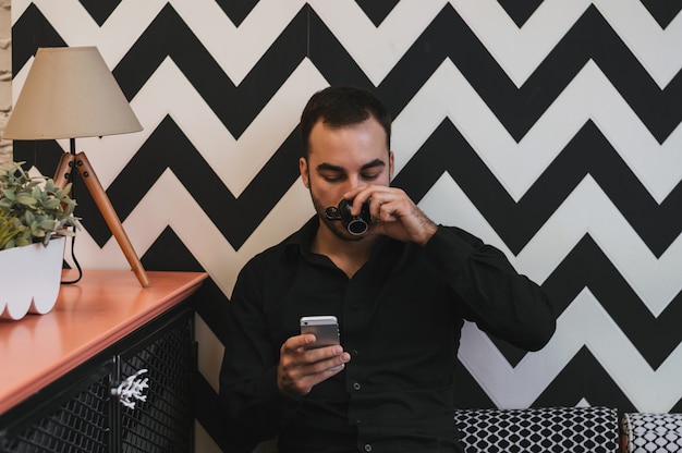 Young guy texting with his mobile phone at the bar and having a cappuccino.