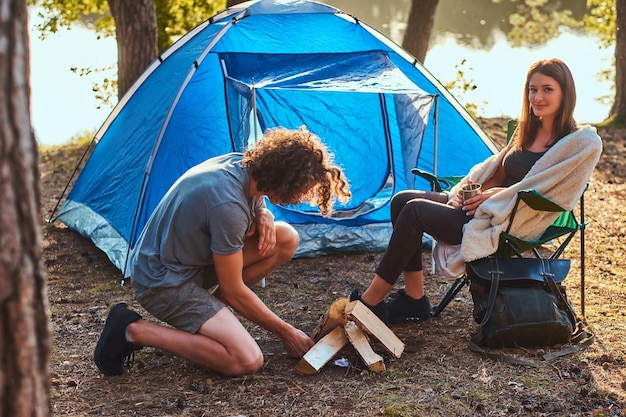 Young guy tries to light a fire. Young hiker couple at camp in the forest. Travel, tourism, and hike concept.