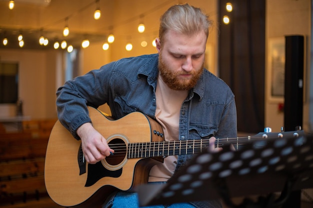 A young guy with a beard plays an acoustic guitar in a room with warm lighting