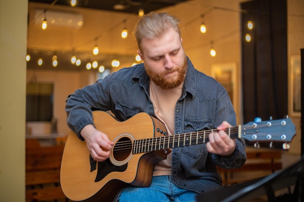 A young guy with a beard plays an acoustic guitar in a room with warm lighting