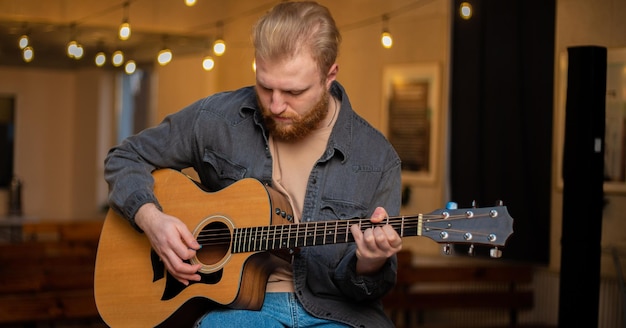 A young guy with a beard plays an acoustic guitar in a room with warm lighting