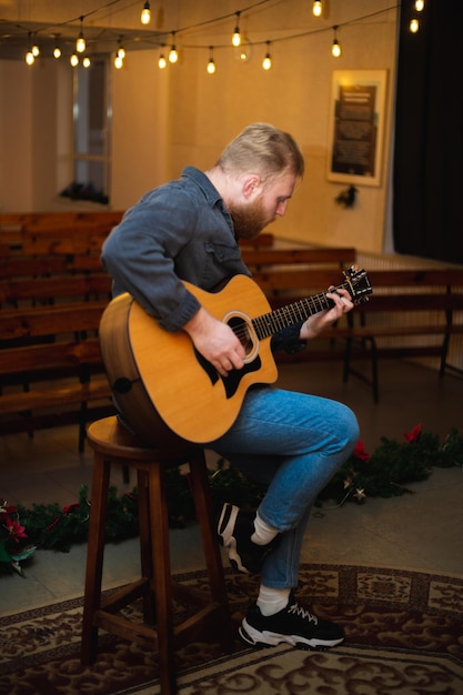 A young guy with a beard plays an acoustic guitar in a room with warm lighting