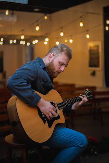 A young guy with a beard plays an acoustic guitar in a room with warm lighting