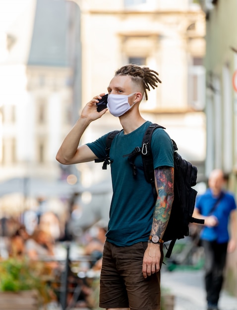 Young guy with face mask and dreadlocks is talking by mobile phone on the street