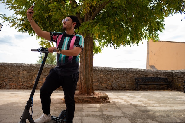 Young guy with glasses and a beard and braids in his hair taking a selfie with his electric scooter