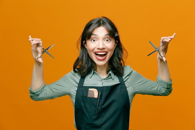 Young hairdresser woman wearing apron holding scissors looking at camera happy and excited smiling broadly standing over orange background