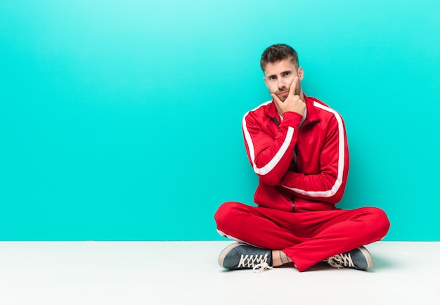 Young handosme man looking serious, thoughtful and distrustful, with one arm crossed and hand on chin, weighting options against flat color wall