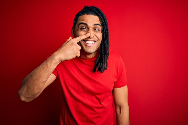 Young handsome african american afro man with dreadlocks wearing red casual tshirt Pointing with hand finger to face and nose smiling cheerful Beauty concept