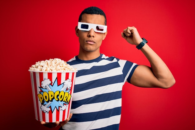 Young handsome african american man watching movie using 3d glasses eating popcorns Strong person showing arm muscle confident and proud of power