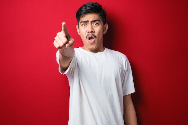 Young handsome asian man wearing casual shirt standing over red background pointing with finger surprised ahead, open mouth amazed expression, something on the front