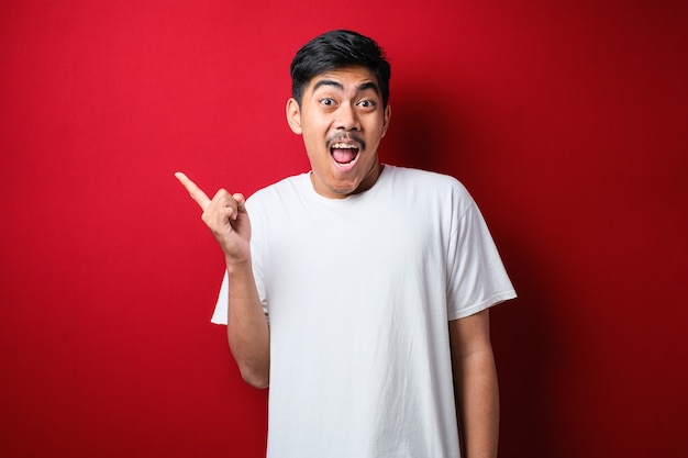 Young handsome asian man wearing white t-shirt over red background with a big smile on face; pointing with hand finger to the side looking at the camera.