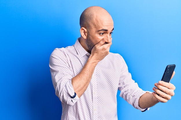 Young handsome bald man using smartphone covering mouth with hand, shocked and afraid for mistake. surprised expression