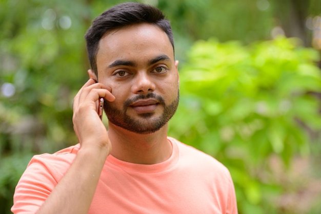 Young handsome bearded Indian man talking on the phone at the park