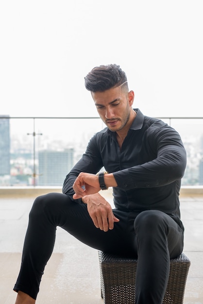 Young handsome bearded Persian businessman sitting and checking the time in the city