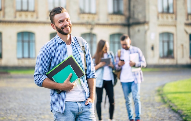 Young handsome bearded student guy in casual clothes with backpack and books in hands is walking and posing on a university  buildings