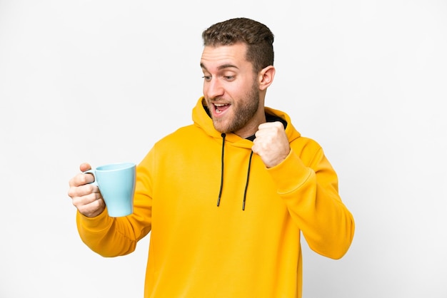 Young handsome blonde man holding cup of coffee over isolated background celebrating a victory