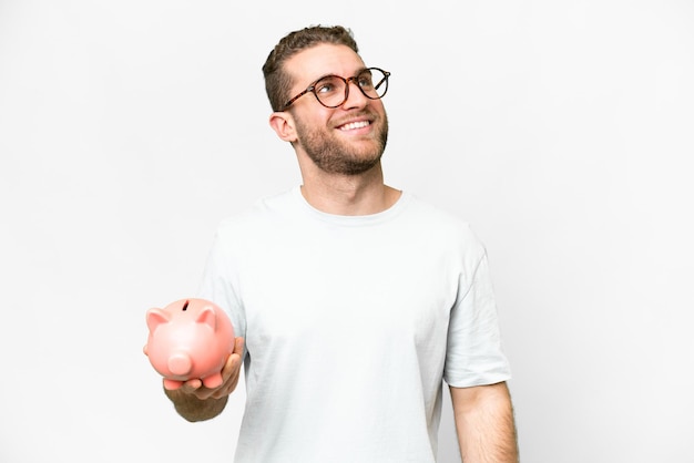 Young handsome blonde man holding a piggybank looking up while smiling