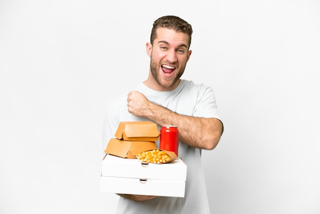 Young handsome blonde man holding pizzas and burgers over isolated background celebrating a victory