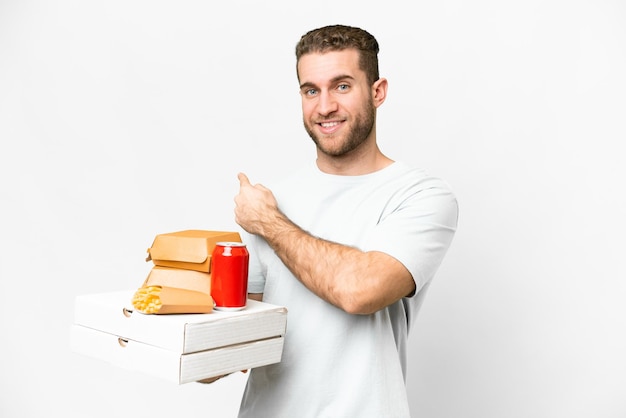 Young handsome blonde man holding pizzas and burgers over isolated background pointing back