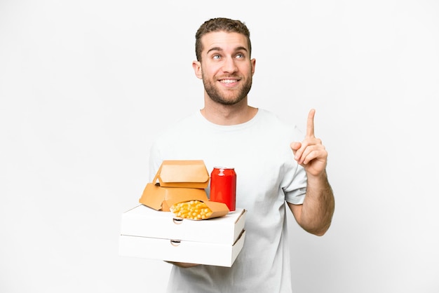 Young handsome blonde man holding pizzas and burgers over isolated background pointing up a great idea