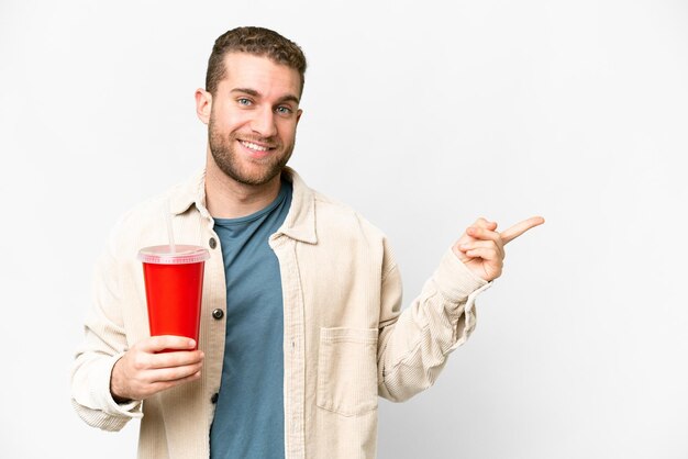 Young handsome blonde man holding soda over isolated white background pointing finger to the side