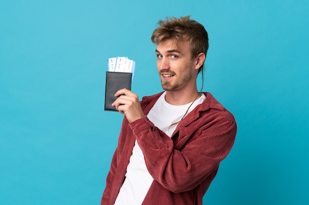 Young handsome blonde man isolated on blue background happy in vacation with passport and plane tickets