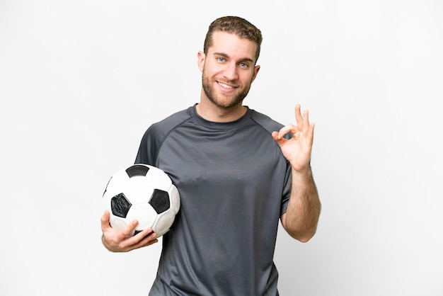 Young handsome blonde man over isolated white background with soccer ball and making OK sign