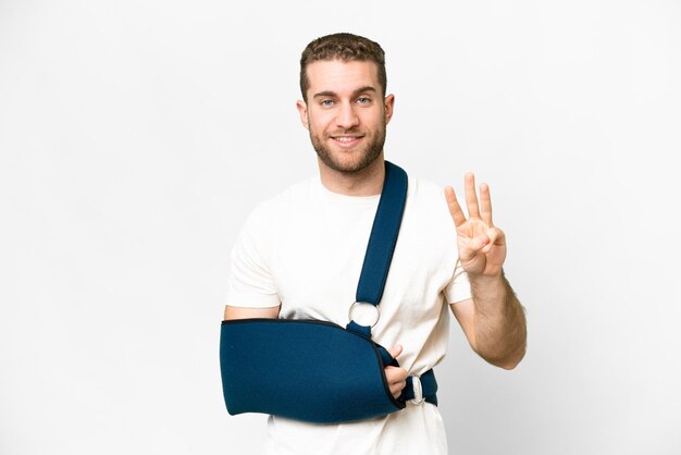 Young handsome blonde man with broken arm and wearing a sling over isolated white background happy and counting three with fingers