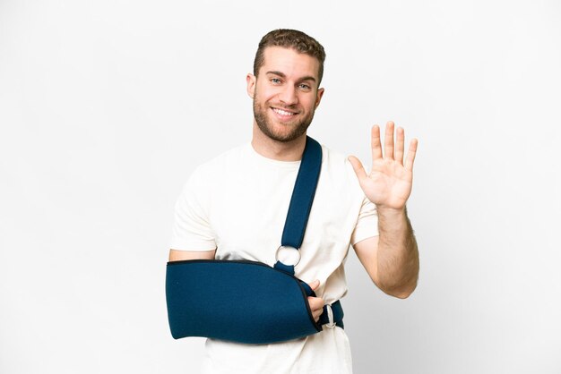 Young handsome blonde man with broken arm and wearing a sling over isolated white background saluting with hand with happy expression