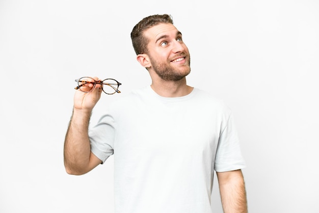 Young handsome blonde man with glasses over isolated white background looking up while smiling