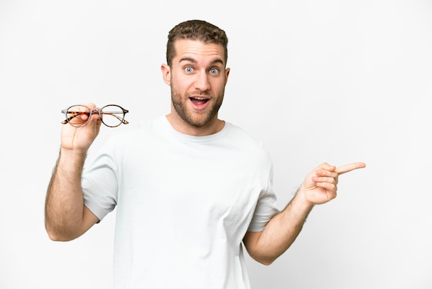 Young handsome blonde man with glasses over isolated white background surprised and pointing finger to the side