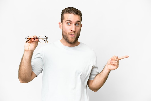 Young handsome blonde man with glasses over isolated white background surprised and pointing side