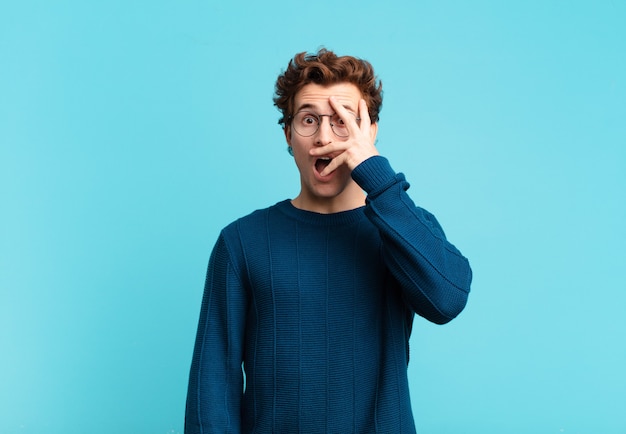 Young handsome boy looking shocked, scared or terrified, covering face with hand and peeking between fingers