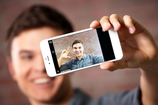 Young handsome boy making photo by his self with mobile phone on brick wall, close up