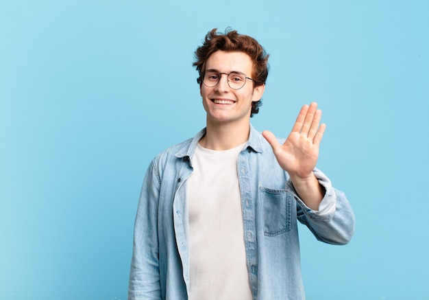 Young handsome boy smiling happily and cheerfully, waving hand, welcoming and greeting you, or saying goodbye