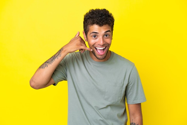 Young handsome Brazilian man isolated on yellow background making phone gesture Call me back sign