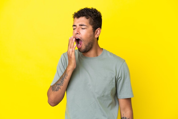 Photo young handsome brazilian man isolated on yellow background yawning and covering wide open mouth with hand