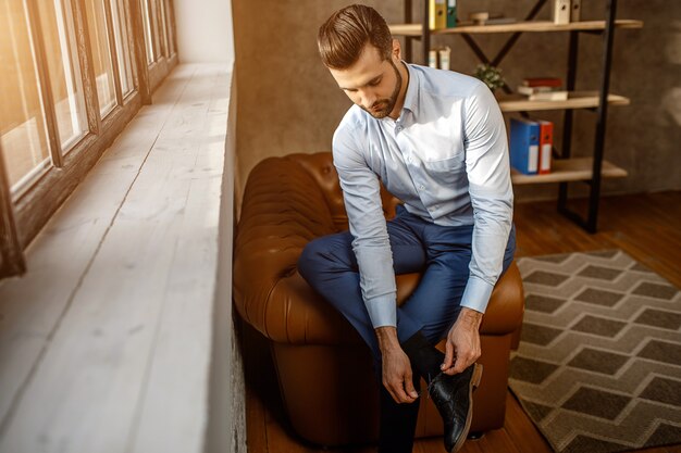 Photo young handsome businessman sit on sofa and tie laces on shoes in his own office. confident guy beside window. sun light shining.