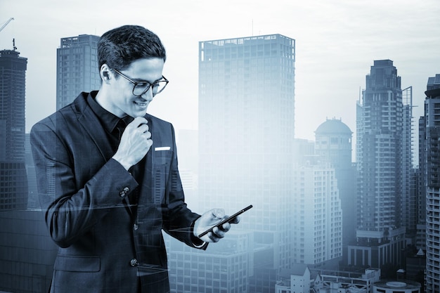 Young handsome businessman in suit and glasses using phone and thinking how to tackle the problem new career opportunities MBA Bangkok on background Double exposure