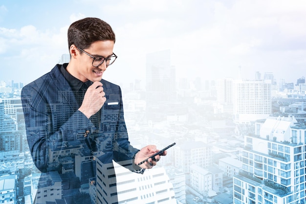 Young handsome businessman in suit and glasses using phone and thinking how to tackle the problem new career opportunities MBA Bangkok on background Double exposure