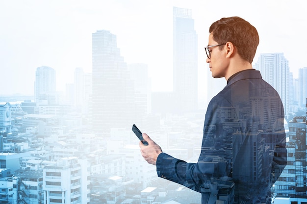 Young handsome businessman in suit and glasses using phone and thinking how to tackle the problem new career opportunities MBA Bangkok on background Double exposure